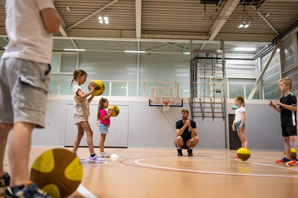 Kinderen volgen een leuke basketbaltraining onder begeleiding van een coach tijdens een basketbalfeestje van VoetbalSkills in een sporthal.